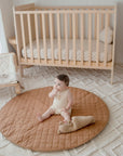 A baby lying on a round brown padded play mat with a quilted design, placed on a rug next to a crib.