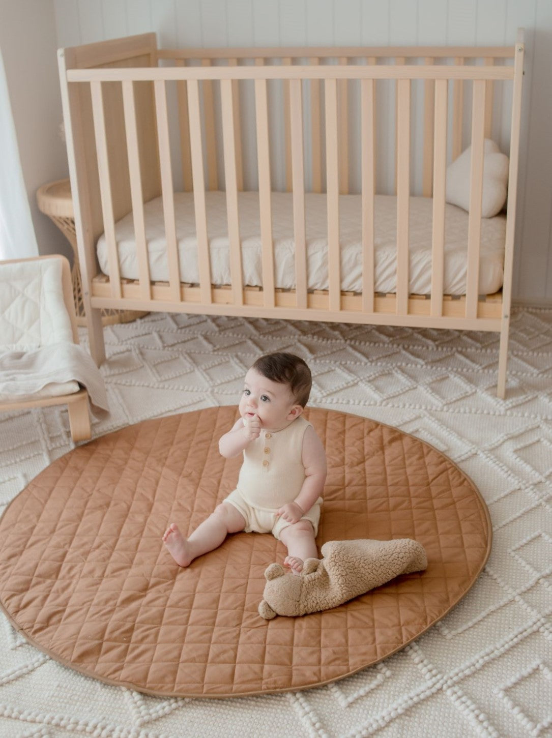A baby lying on a round brown padded play mat with a quilted design, placed on a rug next to a crib.